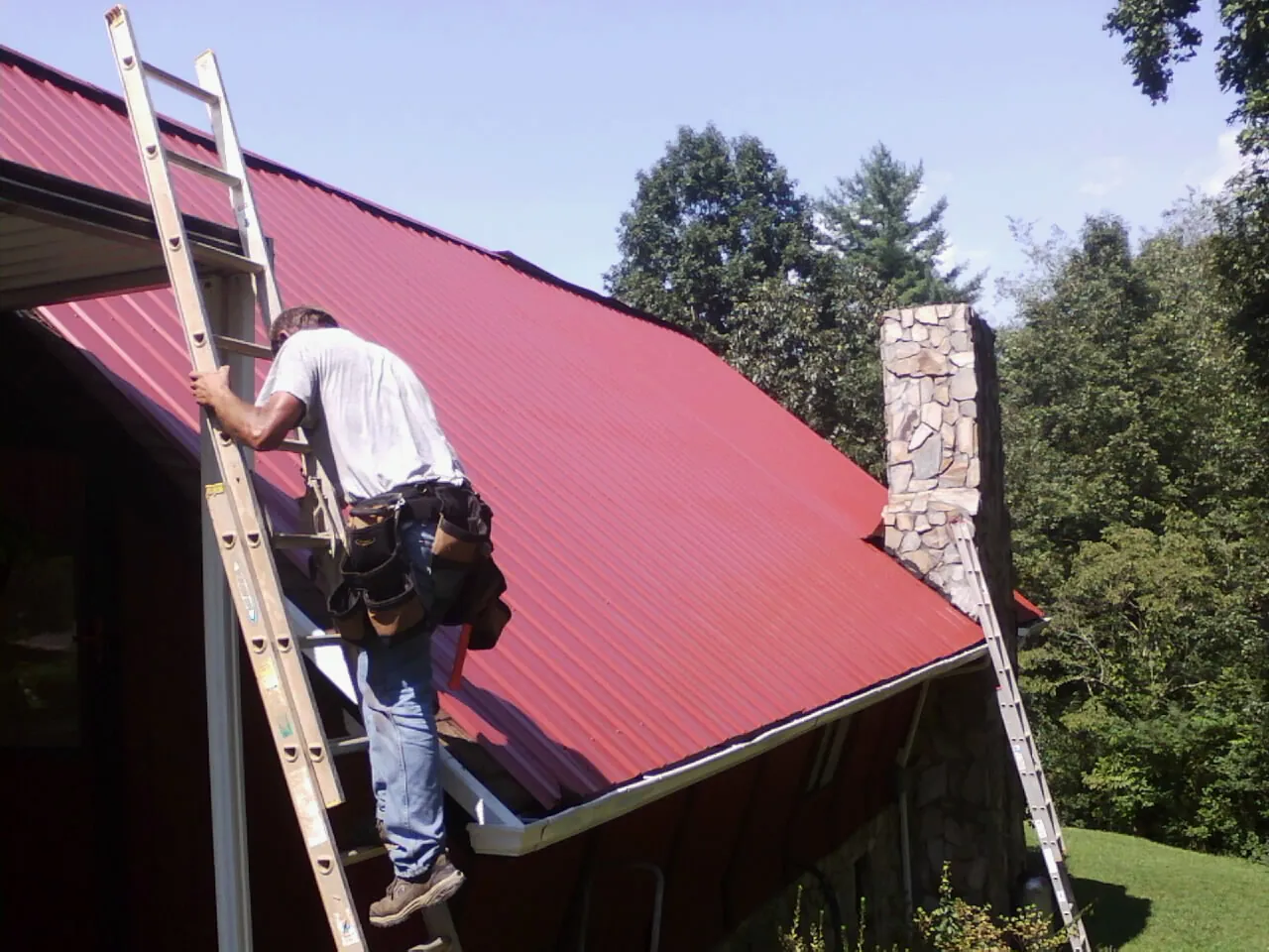 Roofer on a ladder beside a red metal roof and stone chimney — Soffit & Fascia Repair by Roofing Brothers in Wethersfield